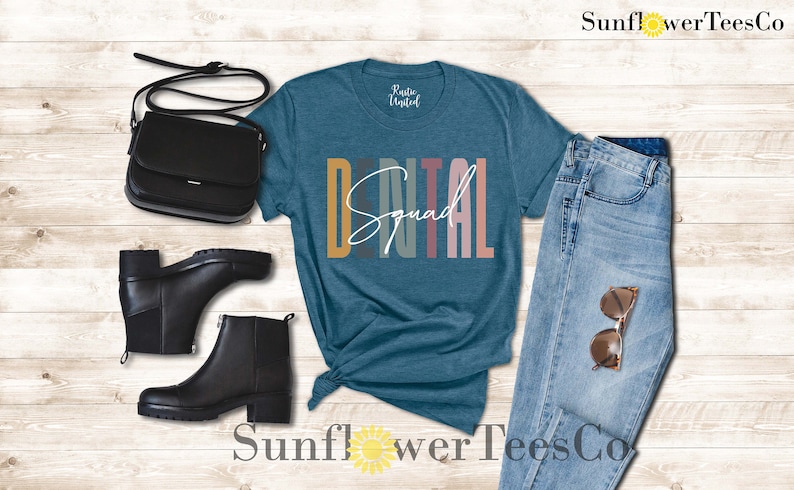 May include: A teal blue t-shirt with the text "Dental Squad" in colorful vertical stripes. The shirt is paired with blue jeans, black boots, and a black purse.