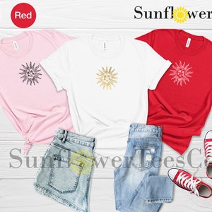 May include: Three t-shirts with a gold sun graphic design. The shirts are pink, white, and red. The shirts are laid out on a wooden surface with a pair of blue jeans and red sneakers.