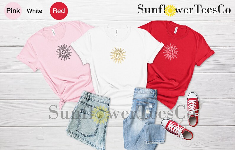 May include: Three t-shirts with a gold sun graphic design. The shirts are pink, white, and red. The shirts are laid out on a wooden surface with a pair of blue jeans and red sneakers.