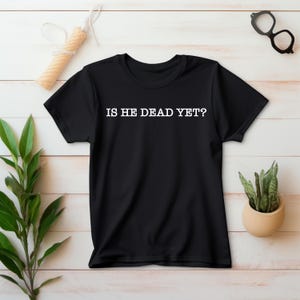 K&ouml;nnte beinhalten: Schwarzes T-Shirt mit wei&szlig;em Text, der "IS HE DEAD YET?" lautet.