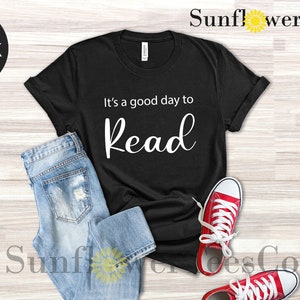 Op de afbeelding: Zwart T-shirt met de tekst "It's a good day to Read" in witte letters. Het shirt wordt gecombineerd met lichtblauwe jeans en rode sneakers met witte veters. De afbeelding staat op een lichte houtnerf achtergrond.