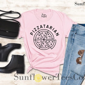 May include: Pink t-shirt with the text "Pizzatarian" and a black and white illustration of a pizza slice. The shirt is paired with blue jeans, black boots, and a black purse.