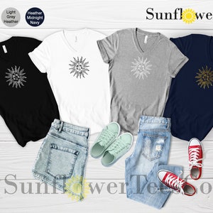 May include: Four V-neck t-shirts with a graphic of a sun on the front. The shirts are in black, white, heather grey, and midnight navy. The shirts are laid out on a wooden surface with a pair of blue jeans and a pair of white sneakers.