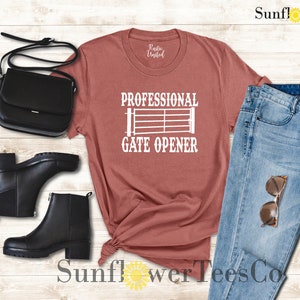 Op de afbeelding: Een roestkleurig T-shirt met de tekst "Professional Gate Opener" en een wit silhouet van een poort. Het shirt wordt gecombineerd met een blauwe jeans, zwarte laarzen en een zwarte tas.