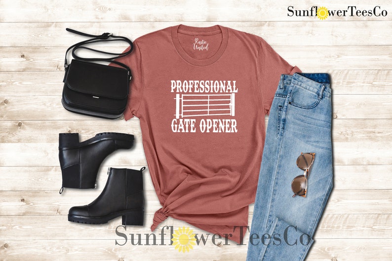 Op de afbeelding: Een roestkleurig T-shirt met de tekst "Professional Gate Opener" en een wit silhouet van een poort. Het shirt wordt gecombineerd met een blauwe jeans, zwarte laarzen en een zwarte tas.