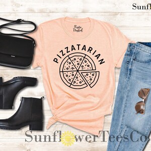 May include: A peach-colored t-shirt with the text "PIZZATARIAN" and a black and white illustration of a pizza slice. The shirt is paired with blue jeans, black boots, and a black purse.