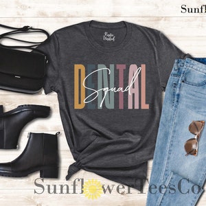 May include: A dark gray t-shirt with the text "DENTAL Squad" in colorful vertical stripes. The shirt is styled with a pair of black boots, a black purse, and blue jeans.