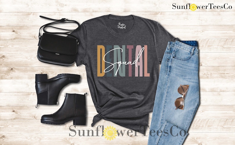 May include: A dark gray t-shirt with the text "DENTAL Squad" in colorful vertical stripes. The shirt is styled with a pair of black boots, a black purse, and blue jeans.