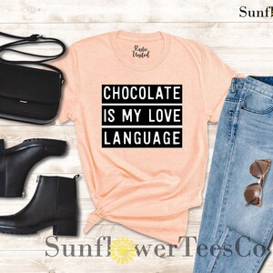 May include: Peach-colored t-shirt with the text "CHOCOLATE IS MY LOVE LANGUAGE" in a black, bold font. The shirt is styled with black ankle boots, a black purse, and blue jeans with sunglasses.