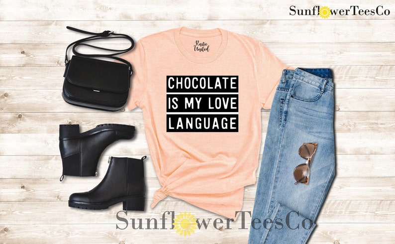 May include: Peach-colored t-shirt with the text "CHOCOLATE IS MY LOVE LANGUAGE" in a black, bold font. The shirt is styled with black ankle boots, a black purse, and blue jeans with sunglasses.