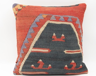 May include: Square decorative pillow with a woven design. The pillow features a red-orange background with a central dark blue geometric pattern. Accents include light beige, red, and purple details. The pillow is likely made of a durable fabric.