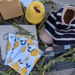 May include: A set of three yellow lemon-patterned fabric wrist cuffs with snaps. The cuffs are on a white background with black leaves. The cuffs are next to a crocheted yellow hat with a brown center. The image also includes a pair of black and white striped baby shoes with a brown leather sole. The shoes have a tag that says "Merci pour votre commande".