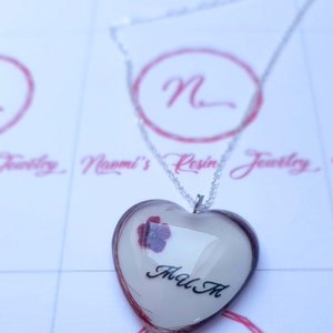 Puede incluir: Collar con colgante de resina en forma de coraz&oacute;n y cadena plateada. El colgante tiene un fondo blanco, una flor morada y las letras "M A M" en negro. Los bordes del coraz&oacute;n est&aacute;n delineados en color marr&oacute;n rojizo.