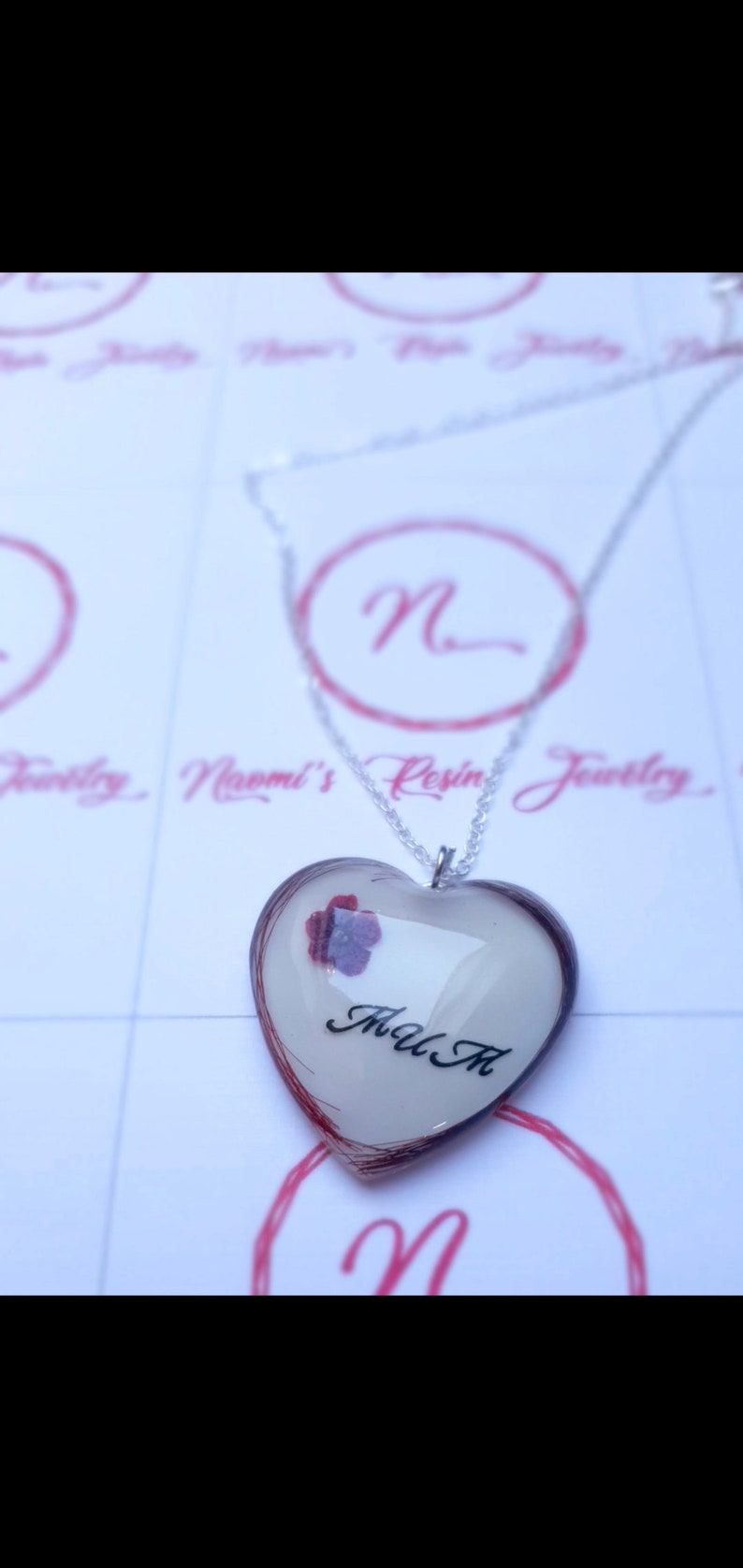 Puede incluir: Collar con colgante de resina en forma de coraz&oacute;n y cadena plateada. El colgante tiene un fondo blanco, una flor morada y las letras "M A M" en negro. Los bordes del coraz&oacute;n est&aacute;n delineados en color marr&oacute;n rojizo.