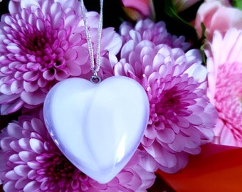 May include: A white heart-shaped pendant hangs from a silver chain against a background of pink and purple flowers.