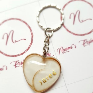Puede incluir: Llavero en forma de coraz&oacute;n con un anillo plateado. El coraz&oacute;n est&aacute; relleno de un material de color claro y mechones de cabello, con el nombre "Jared" en letras doradas. El fondo tiene un logotipo con la letra "N".