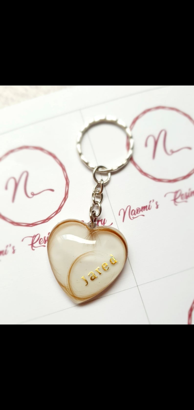 Puede incluir: Llavero en forma de coraz&oacute;n con un anillo plateado. El coraz&oacute;n est&aacute; relleno de un material de color claro y mechones de cabello, con el nombre "Jared" en letras doradas. El fondo tiene un logotipo con la letra "N".