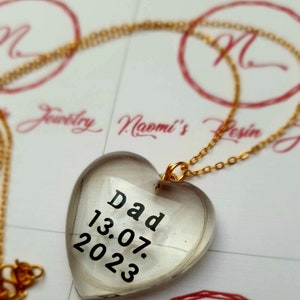 Puede incluir: Collar con colgante en forma de coraz&oacute;n y cadena dorada. El colgante transparente lleva la palabra "Dad" y la fecha "13.07.2023" en negro. El collar est&aacute; sobre una superficie blanca con detalles rojos.