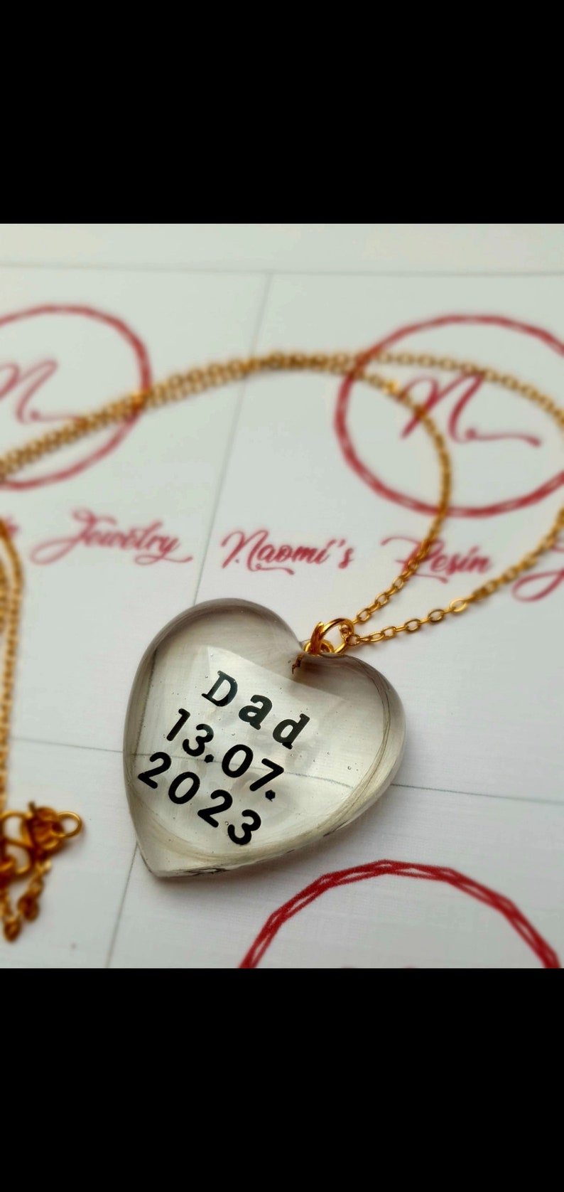 Puede incluir: Collar con colgante en forma de coraz&oacute;n y cadena dorada. El colgante transparente lleva la palabra "Dad" y la fecha "13.07.2023" en negro. El collar est&aacute; sobre una superficie blanca con detalles rojos.