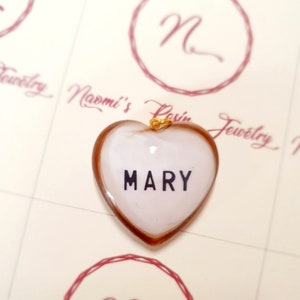 Puede incluir: Colgante en forma de coraz&oacute;n con el nombre "MARY" en letras negras sobre fondo blanco. El colgante tiene una anilla dorada y un borde marr&oacute;n. El fondo presenta un logotipo y las palabras "Naomi's Resin Jewelry".