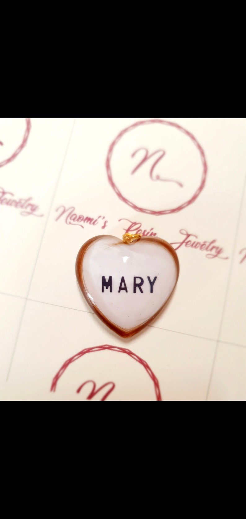 Puede incluir: Colgante en forma de coraz&oacute;n con el nombre "MARY" en letras negras sobre fondo blanco. El colgante tiene una anilla dorada y un borde marr&oacute;n. El fondo presenta un logotipo y las palabras "Naomi's Resin Jewelry".