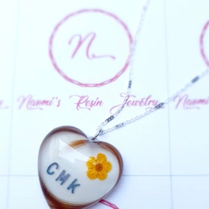 Puede incluir: Collar con colgante de resina en forma de coraz&oacute;n con cadena plateada. El colgante contiene cabello casta&ntilde;o, las iniciales CMK y una peque&ntilde;a flor amarilla. El collar es probablemente hecho a mano.