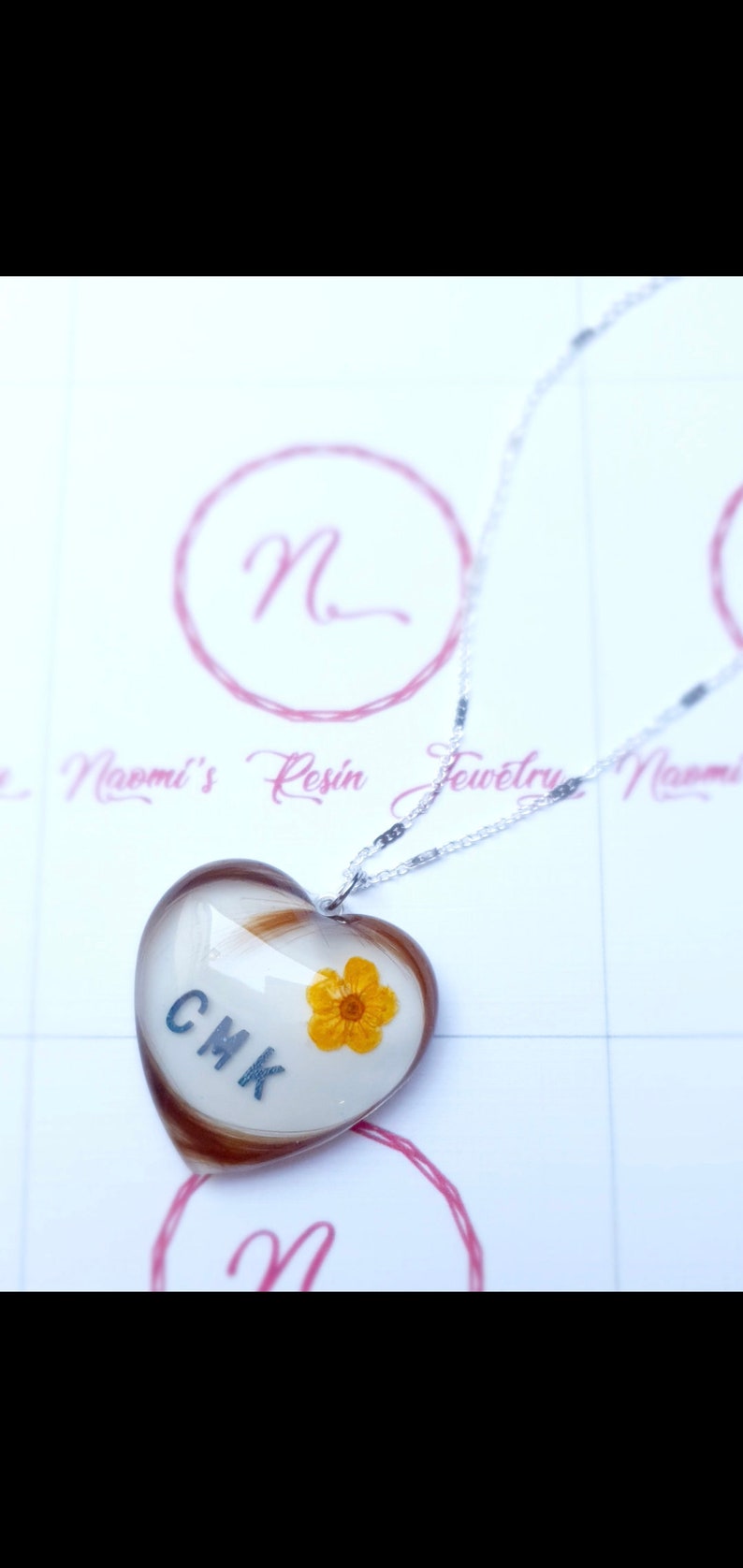 Puede incluir: Collar con colgante de resina en forma de coraz&oacute;n con cadena plateada. El colgante contiene cabello casta&ntilde;o, las iniciales CMK y una peque&ntilde;a flor amarilla. El collar es probablemente hecho a mano.