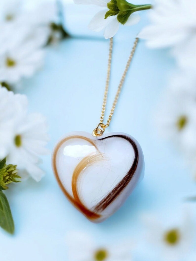 Puede incluir: Un colgante en forma de coraz&oacute;n con una cadena dorada. El colgante es blanco con mechones de cabello casta&ntilde;o y casta&ntilde;o claro. El fondo es azul claro con flores blancas.