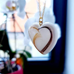 Puede incluir: Un collar con colgante en forma de coraz&oacute;n con una cadena dorada. El colgante transparente contiene mechones de cabello casta&ntilde;o y rubio. El collar cuelga frente a un fondo borroso de flores blancas y una maceta roja.