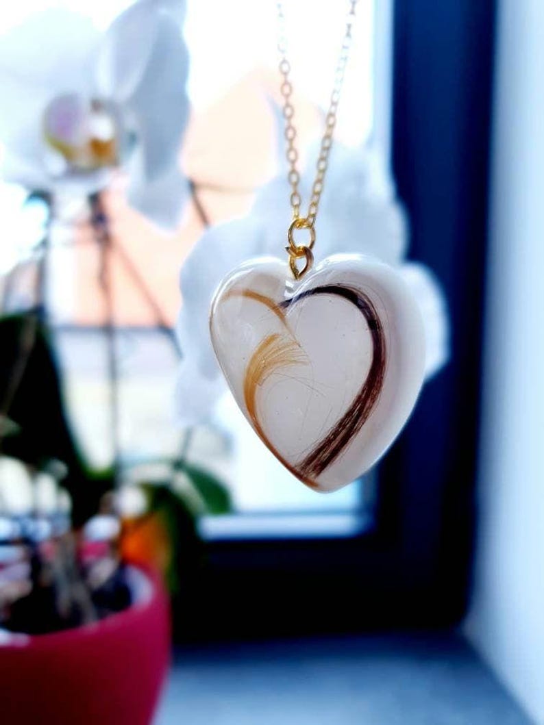 Puede incluir: Un collar con colgante en forma de coraz&oacute;n con una cadena dorada. El colgante transparente contiene mechones de cabello casta&ntilde;o y rubio. El collar cuelga frente a un fondo borroso de flores blancas y una maceta roja.