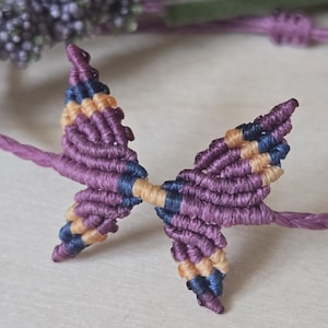 May include: A macrame bracelet featuring a central butterfly motif crafted from purple, dark blue, and tan cord. The butterfly design is connected to a thin, braided purple cord that forms the adjustable band of the bracelet.