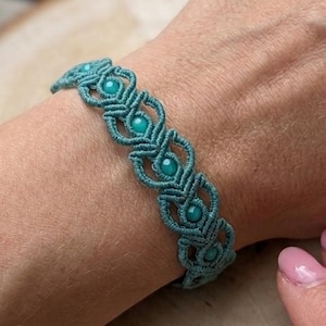 May include: A teal macrame bracelet featuring a series of intricate knotted patterns and small, round, matching teal beads embedded within the center of each decorative loop, creating a continuous, textured design around the wrist.