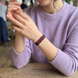 Puede incluir: Una pulsera tejida negra y roja con la palabra "GO" en letras rojas. La pulsera se lleva en la mu&ntilde;eca. La persona lleva un su&eacute;ter de color lila claro. El fondo est&aacute; borroso.
