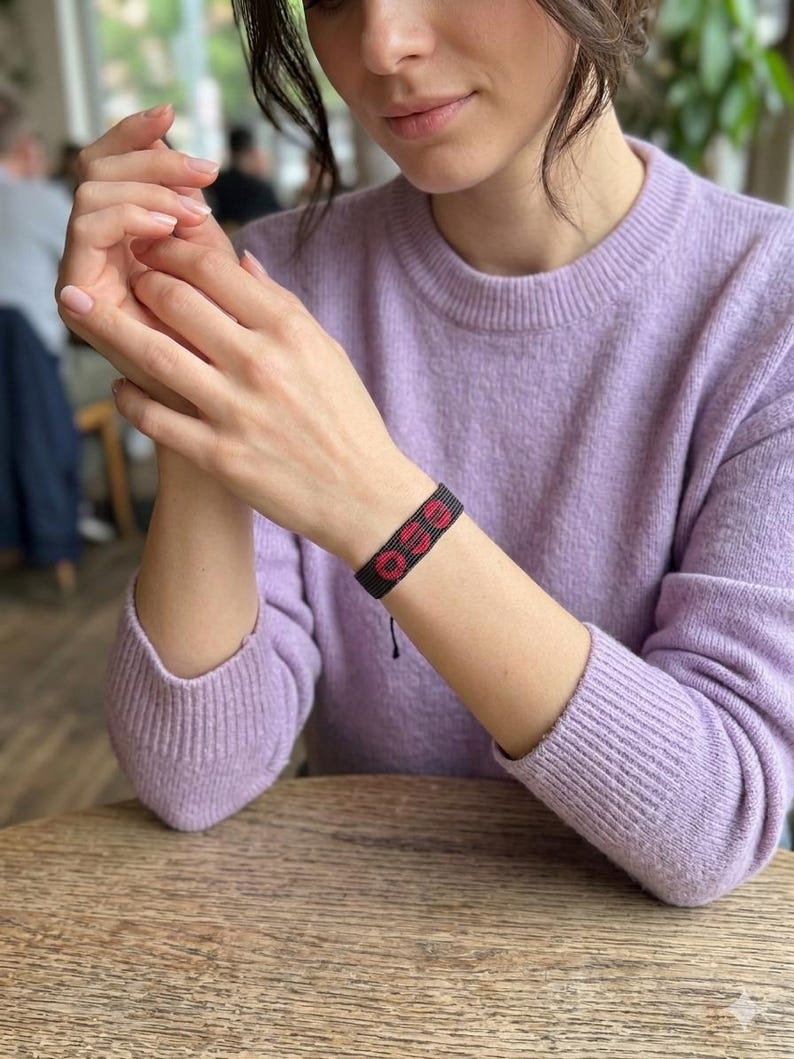 Puede incluir: Una pulsera tejida negra y roja con la palabra "GO" en letras rojas. La pulsera se lleva en la mu&ntilde;eca. La persona lleva un su&eacute;ter de color lila claro. El fondo est&aacute; borroso.
