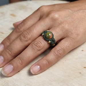 May include: A ring featuring a central round, mottled green and orange stone set within dark green knotted cord. The band is accented with small, metallic gold-toned beads integrated into the intricate macrame-style fiber design.