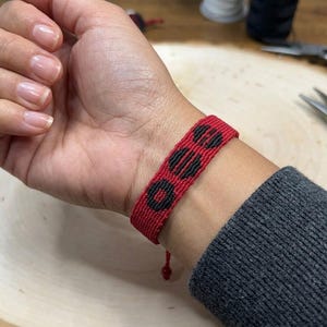 Puede incluir: Pulsera tejida roja y negra con un patr&oacute;n geom&eacute;trico de tres c&iacute;rculos negros. La pulsera se lleva en la mu&ntilde;eca y el fondo incluye herramientas y otros suministros de artesan&iacute;a. La pulsera mide aproximadamente 2 cm de ancho.