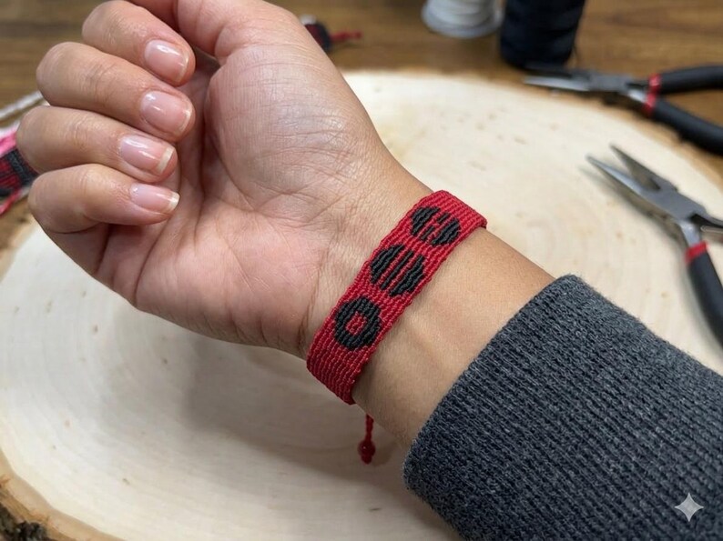 Puede incluir: Pulsera tejida roja y negra con un patr&oacute;n geom&eacute;trico de tres c&iacute;rculos negros. La pulsera se lleva en la mu&ntilde;eca y el fondo incluye herramientas y otros suministros de artesan&iacute;a. La pulsera mide aproximadamente 2 cm de ancho.