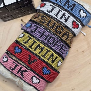 May include: A collection of colorful woven friendship bracelets. Each bracelet features a name in black letters with a heart symbol. The bracelets are in various colors, including blue, white, brown, purple, yellow, red, and pink. A Rubik's cube is in the background.