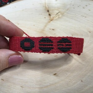 Puede incluir: Una pulsera de macram&eacute; roja con tres dise&ntilde;os circulares negros. La pulsera, sostenida por una mano, muestra un intrincado patr&oacute;n tejido y un color vibrante. Un accesorio hecho a mano.