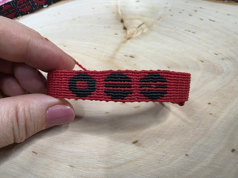 Puede incluir: Una pulsera de macram&eacute; roja con tres dise&ntilde;os circulares negros. La pulsera, sostenida por una mano, muestra un intrincado patr&oacute;n tejido y un color vibrante. Un accesorio hecho a mano.
