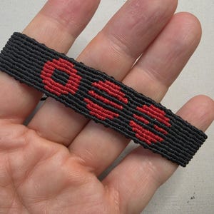 Puede incluir: Pulsera tejida negra con la palabra "OSS" en hilo rojo. La pulsera es rectangular y parece hecha a mano. Las letras son en negrita y est&aacute;n centradas en el fondo negro.