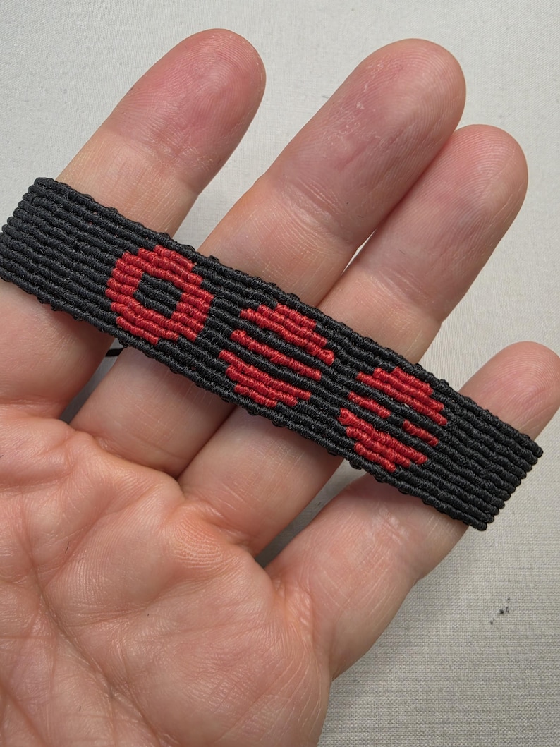 Puede incluir: Pulsera tejida negra con la palabra "OSS" en hilo rojo. La pulsera es rectangular y parece hecha a mano. Las letras son en negrita y est&aacute;n centradas en el fondo negro.