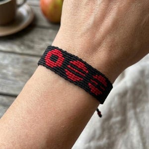 Puede incluir: Pulsera tejida negra y roja con un patr&oacute;n repetitivo. La pulsera se lleva en la mu&ntilde;eca. La pulsera est&aacute; hecha de un material tejido. La pulsera es un art&iacute;culo hecho a mano.