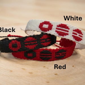 Puede incluir: Tres pulseras de amistad tejidas en negro, rojo y blanco. Cada pulsera presenta un patr&oacute;n repetitivo de c&iacute;rculos rojos. Las pulseras est&aacute;n apiladas sobre una superficie de madera. Las palabras "White", "Black" y "Red" est&aacute;n escritas en la imagen.