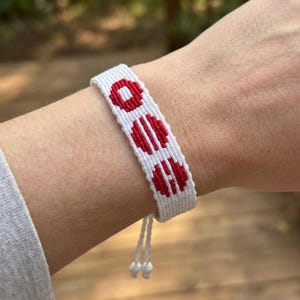Puede incluir: Pulsera blanca tejida con tres dise&ntilde;os circulares rojos. La pulsera est&aacute; en una mu&ntilde;eca y tiene cordones blancos ajustables con peque&ntilde;as cuentas blancas. El fondo est&aacute; borroso con follaje verde.
