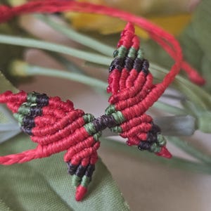 May include: A red macrame butterfly bracelet with green and black accents. The butterfly is the focal point, with a detailed woven design. The bracelet is set against a backdrop of green leaves and flowers, creating a natural aesthetic.