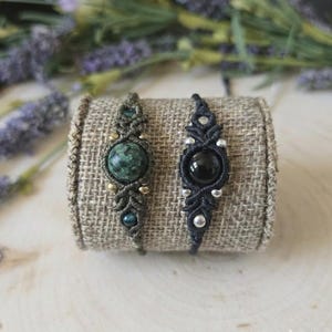 May include: Two macrame bracelets, one olive green with a green stone and the other black with a black stone. Both bracelets are displayed on a tan fabric roll, with lavender sprigs in the background.