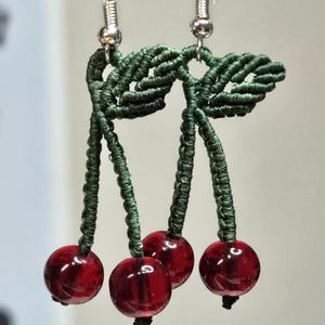 May include: A pair of handmade cherry earrings. The earrings feature three red glass beads that resemble cherries, suspended from green macrame stems and leaves. The ear wires are silver-colored.