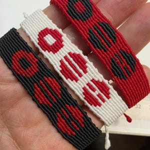 Puede incluir: Tres pulseras de amistad hechas a mano en rojo, negro y blanco. Cada pulsera presenta un patr&oacute;n repetitivo de formas geom&eacute;tricas rojas y negras. Las pulseras est&aacute;n hechas de hilo tejido y tienen un cierre simple anudado.