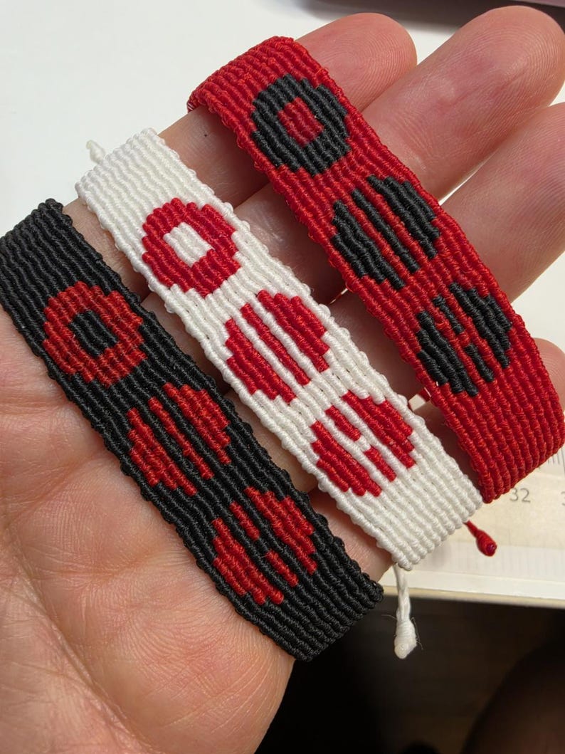 Puede incluir: Tres pulseras de amistad hechas a mano en rojo, negro y blanco. Cada pulsera presenta un patr&oacute;n repetitivo de formas geom&eacute;tricas rojas y negras. Las pulseras est&aacute;n hechas de hilo tejido y tienen un cierre simple anudado.